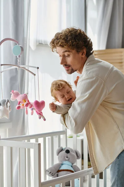 Caring single father holding his crying infant son near baby crib in nursery, fatherhood and love — Stock Photo