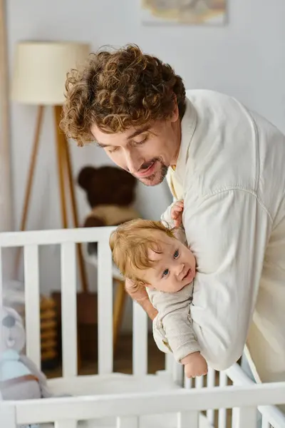 Aimant père bouclé soulevant son fils bébé avec les yeux bleus de la crèche de bébé dans la pépinière, la paternité — Photo de stock