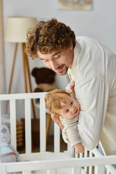 Loving curly father lifting his infant son with blue eyes from baby crib in nursery, fatherhood — Stock Photo