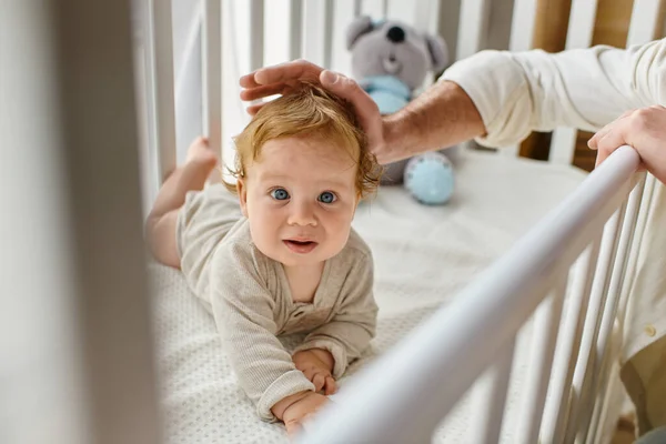 Vue recadrée de l'homme caressant les cheveux de bébé garçon aux yeux bleus dans la crèche, main du père près de l'enfant — Photo de stock