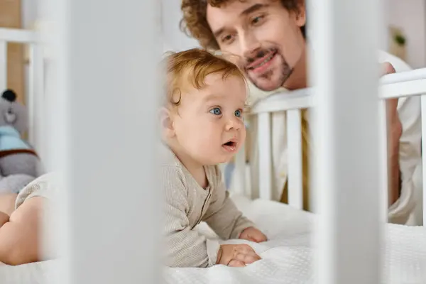 Cute infant baby boy with blue eyes lying in crib while father watching him, man on blurred backdrop — Stock Photo