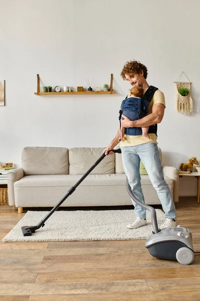 Happy man with infant baby boy in carrier vacuuming apartment at home, father and son bonding — Stock Photo