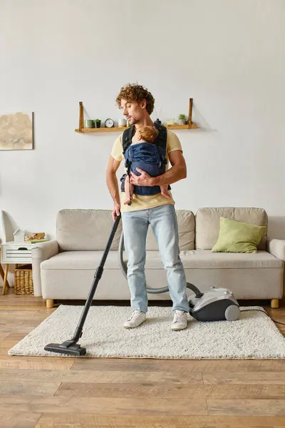 Father with infant baby boy in carrier vacuuming apartment at home, cleanliness and housework — Stock Photo