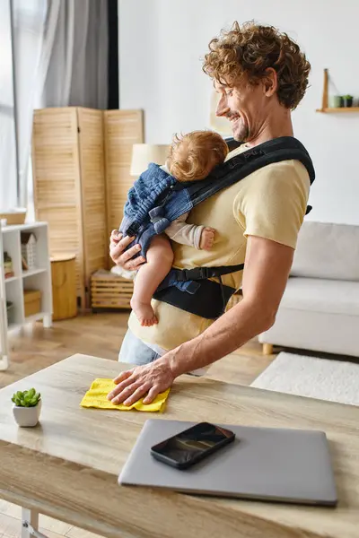 Happy father with infant son in carrier wiping table with yellow rag near gadgets and tiny plant — Stock Photo