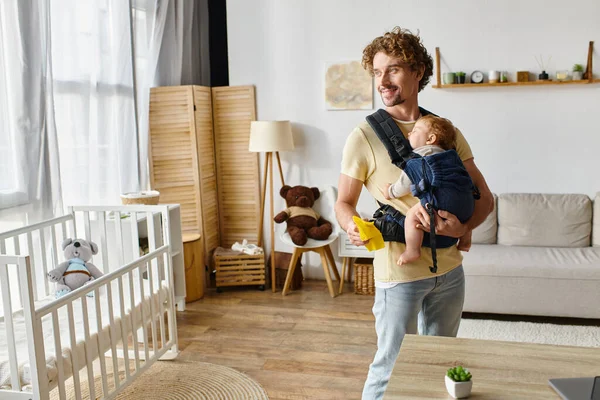 Happy father with infant son in carrier holding yellow rag near working desk in modern apartment — Stock Photo