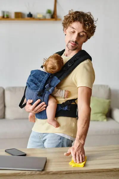 Rizado hombre con bebé hijo en portador limpiar mesa de trabajo con trapo amarillo cerca de gadgets, tareas domésticas - foto de stock