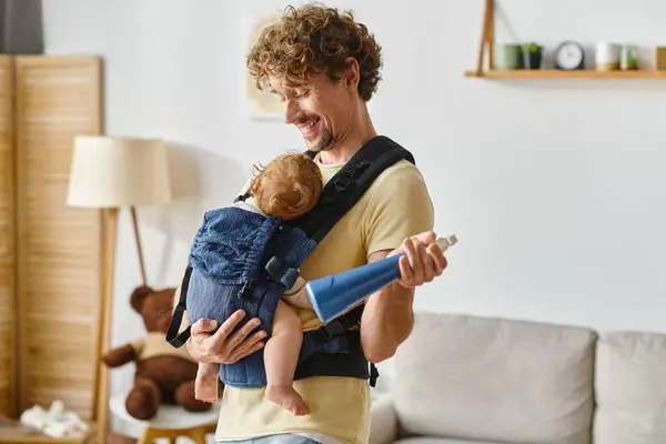 Homme heureux regardant son fils en bas âge dans le porte-bébé tout en tenant le flacon de pulvérisation et chiffon jaune, sourire — Photo de stock