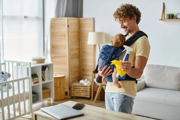 Curly man with infant son in carrier holding spray bottle and yellow rag near gadgets, housework — Stock Photo