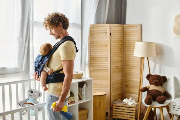 Hombre feliz con hijo pequeño en portador sosteniendo botella de spray y trapo amarillo cerca de cuna, tareas domésticas - foto de stock