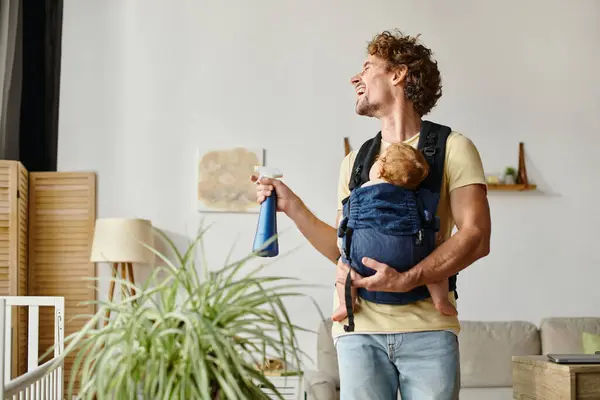 Happy father with infant son in carrier holding spray bottle while laughing and watering green plant — Stock Photo