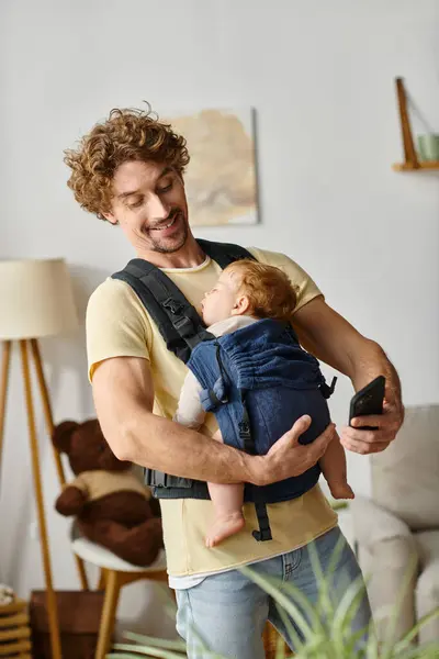 Cheveux bouclés et heureux message de l'homme sur smartphone pendant que bébé dort dans le transporteur, parentalité moderne — Photo de stock