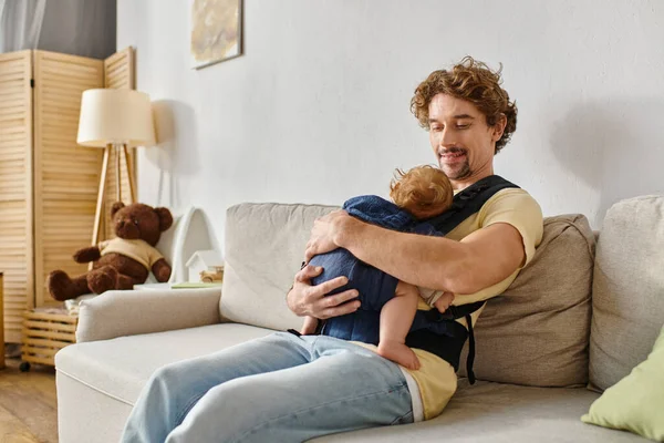 Feliz padre de pelo rizado sentado en el sofá con el niño en el portabebés, la paternidad y el amor - foto de stock