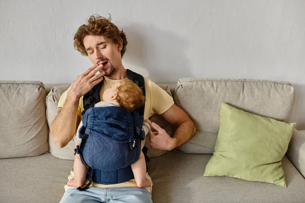 Tired curly haired man yawning with infant son in baby carrier in living room, fatherhood and love — Stock Photo
