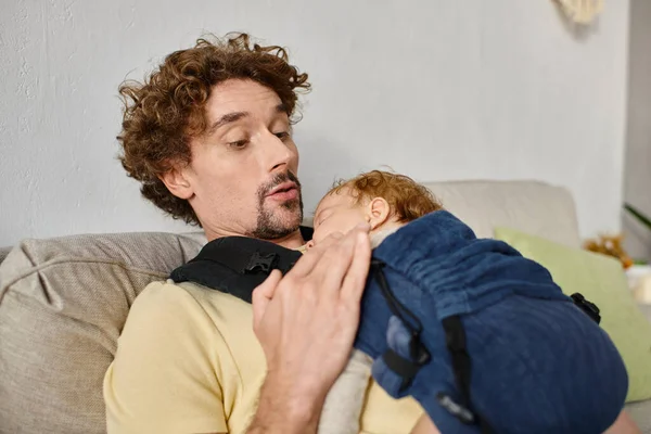 Homem espantado com cabelo encaracolado olhando para seu filho bebê em portador de bebê, paternidade e amor — Fotografia de Stock