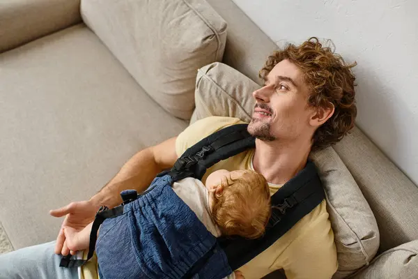 Vista superior del padre feliz sentado en el sofá con el niño en el portabebés, la paternidad y el amor - foto de stock