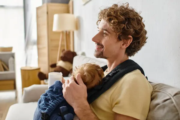 Padre complacido con hijo pequeño en portabebés sosteniendo taza de té en la sala de estar, paternidad - foto de stock