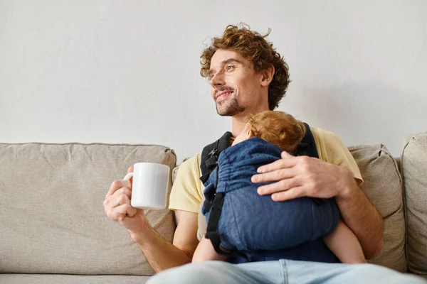 Cheerful father with infant son in baby carrier holding cup of tea in living room, fatherhood — Stock Photo