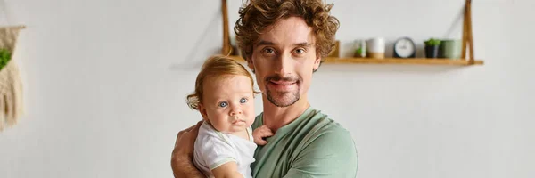Padre de pelo rizado con barba sosteniendo a su hijo pequeño con ojos azules en una acogedora sala de estar, pancarta - foto de stock