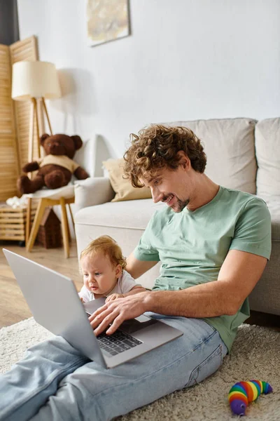 Padre felice utilizzando laptop vicino figlio neonato in soggiorno, equilibrio tra paternità e lavoro — Foto stock