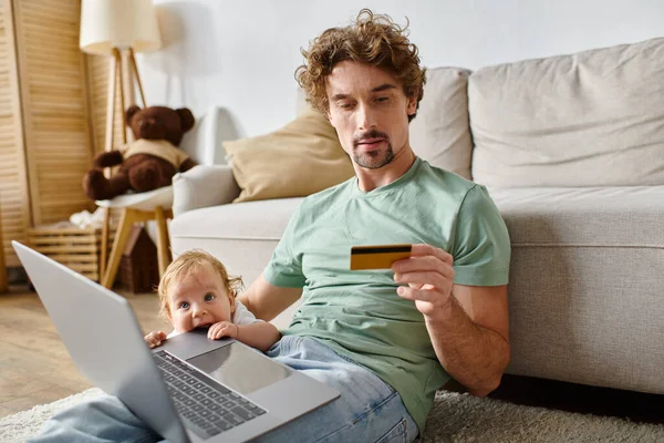 Hombre con pelo rizado celebración de la tarjeta de crédito, mientras que las compras en línea cerca del niño lindo en la sala de estar - foto de stock