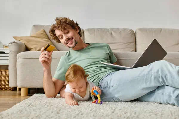 Веселий батько тримає кредитну картку під час онлайн-покупки біля дитячого хлопчика, який грає з биком — стокове фото
