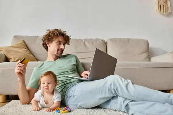 Hombre alegre sosteniendo la tarjeta de crédito mientras que hace compras en línea cerca del niño en la alfombra con el sonajero - foto de stock