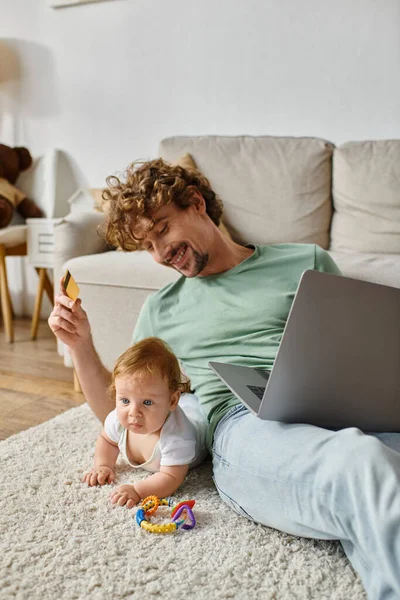 Sonriente hombre sosteniendo tarjeta de crédito mientras hace compras en línea cerca de bebé niño en la alfombra con sonajero - foto de stock
