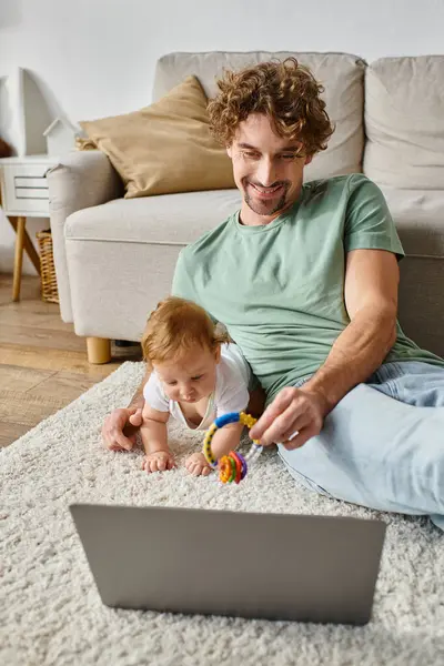 Glücklicher Mann hält Babyrassel neben Säugling und Laptop auf Teppich und balanciert zwischen Arbeit und Leben — Stockfoto