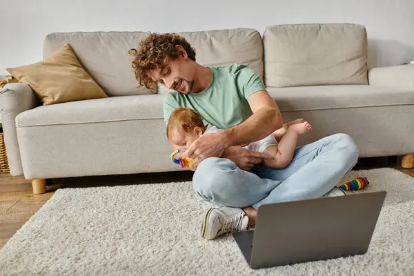 Glücklicher Mann mit lockigem Haar hält Babyrassel in der Nähe von Säugling und Laptop, Work-Life-Balance — Stockfoto