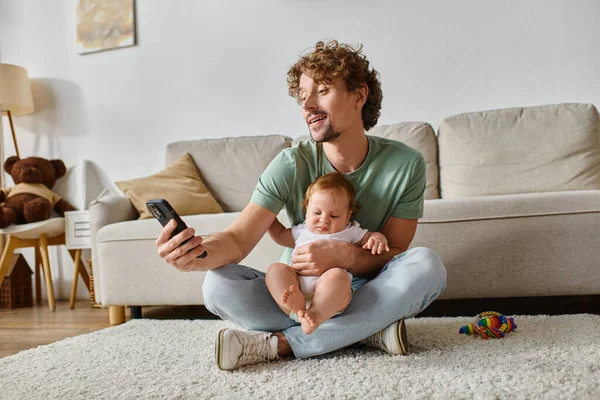 Happy single father using smartphone while sitting with infant baby boy on carpet near rattle — Stock Photo