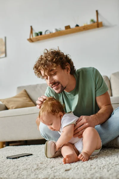 Feliz padre soltero usando el teléfono inteligente mientras está sentado con bebé niño en la alfombra en casa - foto de stock