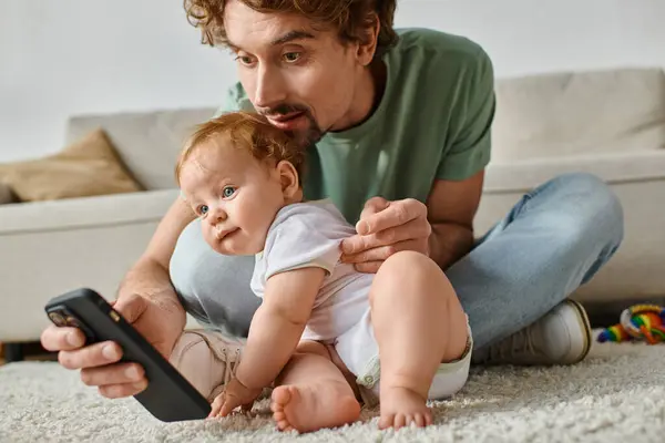 Cheerful single father using smartphone while sitting with infant baby boy on carpet at home — Stock Photo