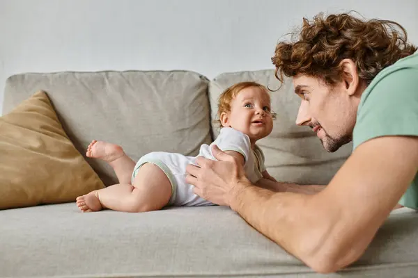 Отец смотрит на милого младенца-сына и весело проводит время на диване в гостиной, прощаясь — стоковое фото
