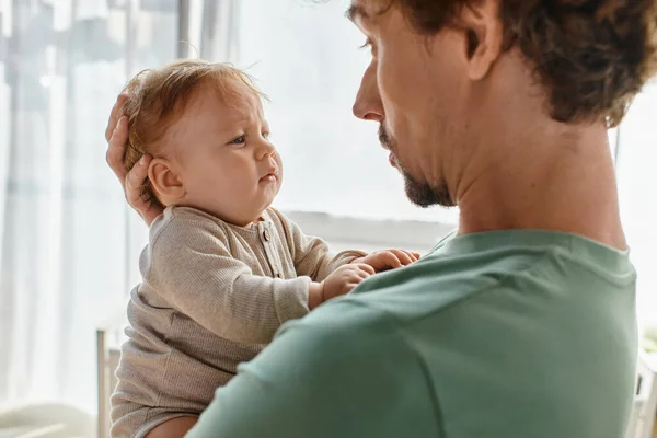 Père attentionné avec les cheveux bouclés et la barbe tenant dans les bras son petit garçon en vêtements de bébé, portrait — Photo de stock