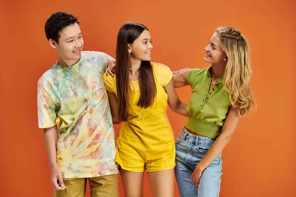 Tres adolescentes alegres diversos en trajes casuales vibrantes mirándose, día de la amistad - foto de stock