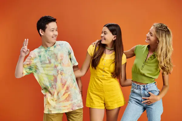 Alegres adolescentes multiculturales en trajes casuales vívidos mirándose el uno al otro en el fondo naranja - foto de stock