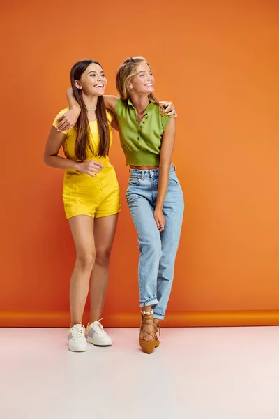 Alegres meninas adolescentes bonitas em roupas vibrantes abraçando e olhando para o fundo laranja — Fotografia de Stock