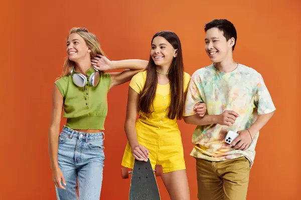 Alegre interracial adolescentes con auriculares y monopatín mirando hacia otro lado en naranja fondo - foto de stock