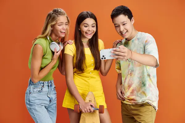 Jolly adolescent diverse friends with headphones and skateboard taking selfie on orange backdrop — Stock Photo