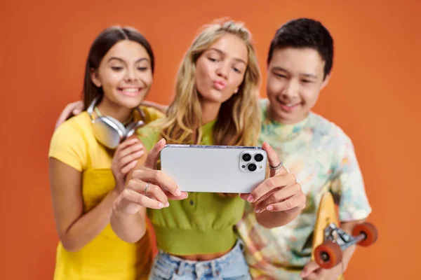 Focus sur smartphone dans les mains de trois amis divers joyeux flous prenant selfie sur fond orange — Photo de stock