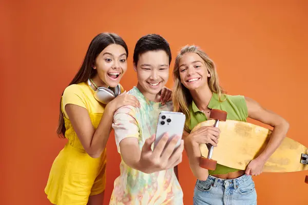 Cheerful multiracial teenage friends in vivid attires with headphones and skateboard taking selfie — Stock Photo