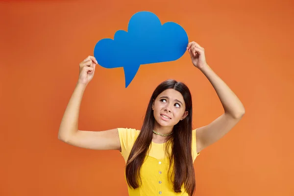 Confused teenage girl in casual attire holding thought bubble above her head on orange backdrop — Stock Photo