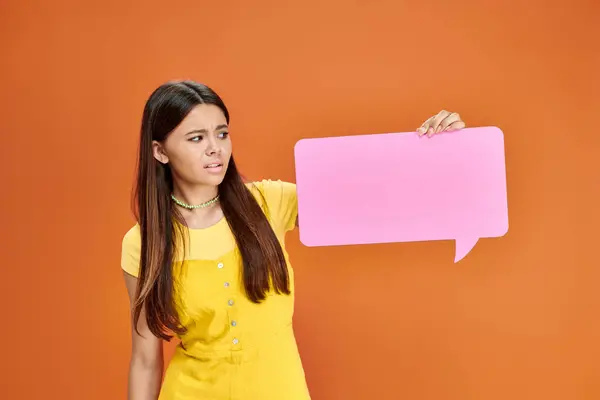 Confusa chica bastante adolescente en traje vibrante sosteniendo burbuja de habla rosa sobre fondo naranja - foto de stock