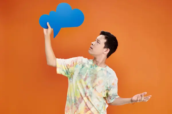 Confused adolescent asian boy in everyday attire looking at blue thought bubble on orange backdrop — Stock Photo