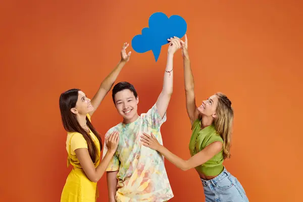 Joyous teenage girls reaching blue thought bubble in hands of asian cheerful boy on orange backdrop — Stock Photo