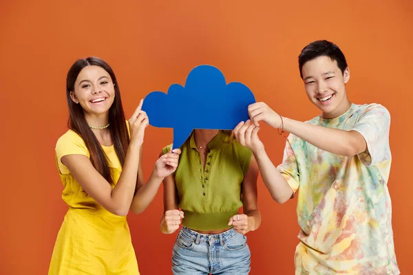 Joyous teenage asian boy and brunette girl holding thought bubble in front of their friend — Stock Photo