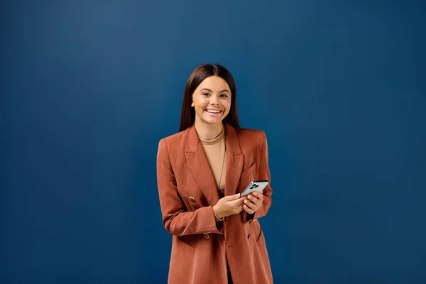 Cheerful fashionable teenage girl in brown blazer holding smartphone and smiling happily at camera — Stock Photo