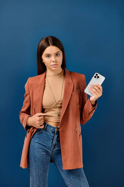 Підліткова стильна дівчина в елегантному коричневому блістері позує з мобільним телефоном в руці і дивиться на камеру — стокове фото
