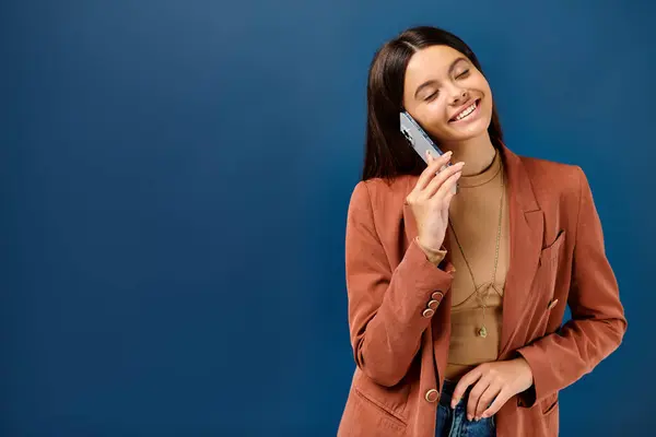 Cheerful trendy adolescent girl in brown blazer talking by phone happily on dark blue background — Stock Photo