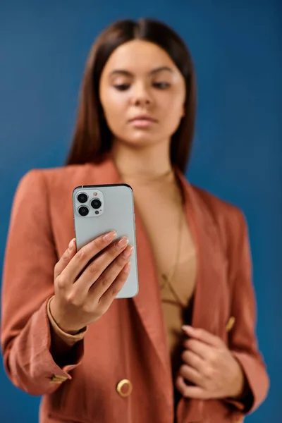 Centrarse en el teléfono inteligente en manos de la chica adolescente borrosa con estilo en chaqueta marrón sobre fondo azul oscuro - foto de stock